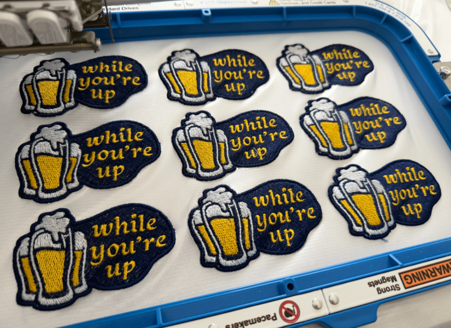 A machine tray filled with multiple embroidered while you're up patches in navy, gold, and white.